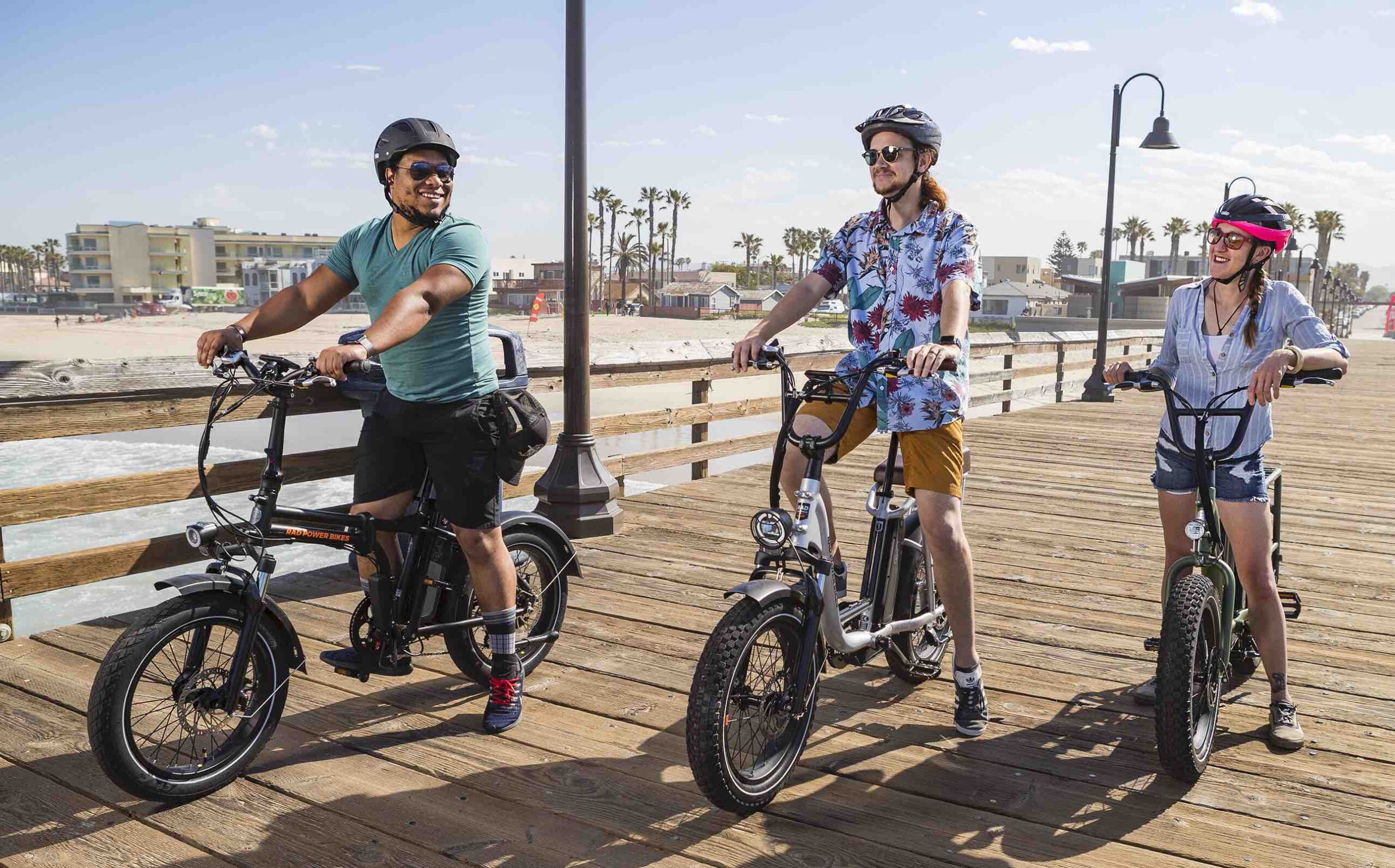 3 friends ride electric bikes down a sunny California boardwalks while laughing.