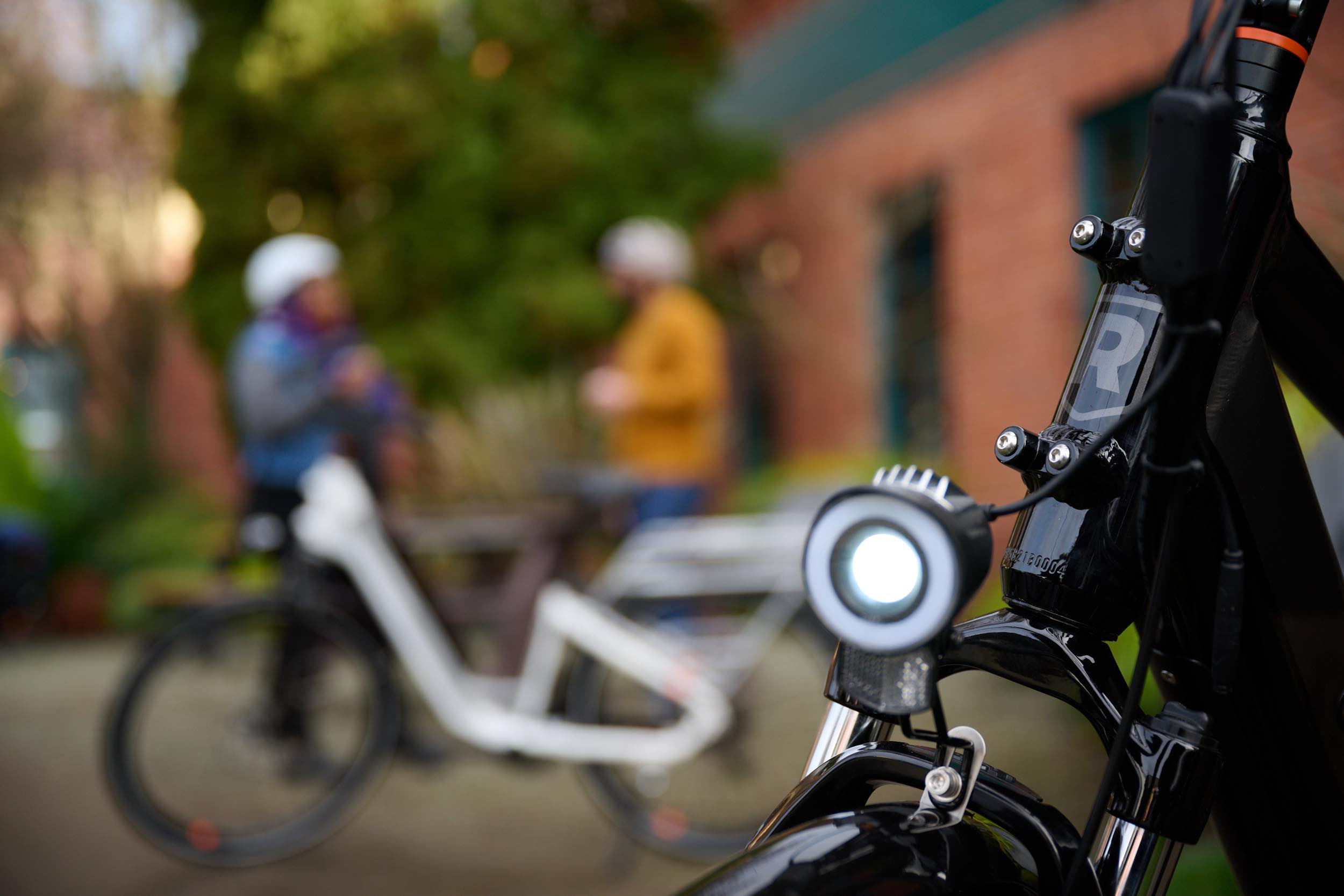 A close-up of an ebike headlight in the rain. In the background, a man and woman chat alongside their bikes.