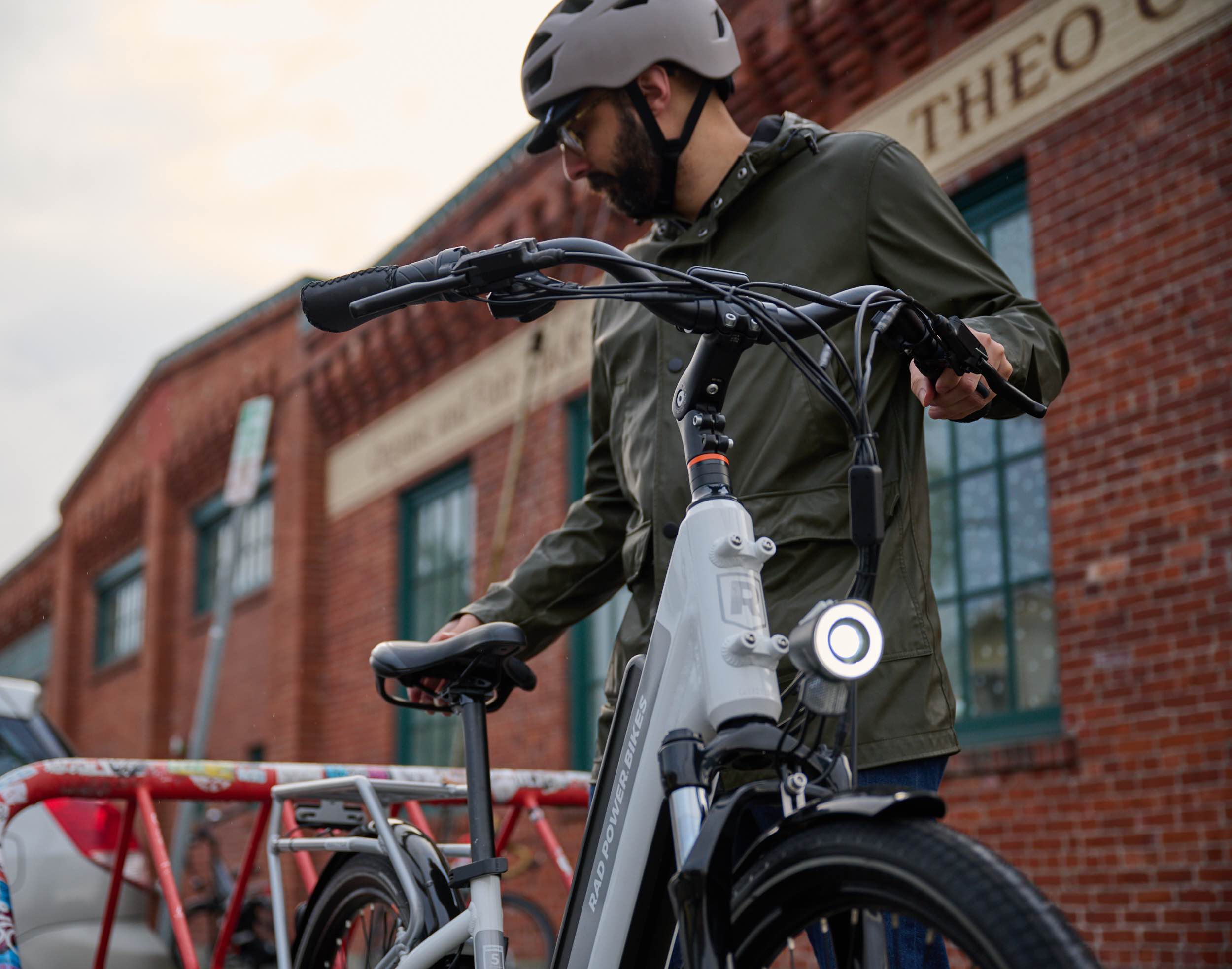 A man in a green coat parks his ebike outside by a bike rack. A red brick building is in the background.